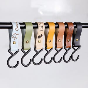 May include: A row of seven leather hooks in various colors, each with a black metal hook. The hooks are attached to a black metal bar. Each hook has a name and a small graphic. The colors include light blue, green, beige, yellow, brown, and black.