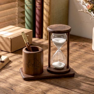 May include: A wooden hourglass with white sand, a pen holder, and a pen. The hourglass is set on a wooden base, and the pen holder has an engraved design. The pen is gold and brown. The items are on a wooden surface.