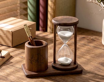 May include: A wooden hourglass with white sand, a pen holder, and a pen. The hourglass is set on a wooden base, and the pen holder has an engraved design. The pen is gold and brown. The items are on a wooden surface.