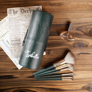 May include: A dark green cylindrical makeup brush holder with the name "Isabella" in white script. The holder is next to a set of makeup brushes, a perfume bottle, and a vintage newspaper on a wooden surface.