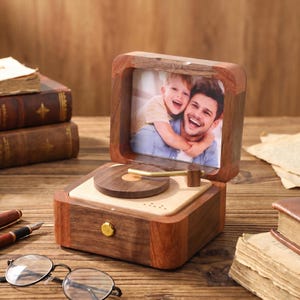 K&ouml;nnte beinhalten: Eine Holz-Spieluhr in Form eines Vintage-Plattenspielers mit einem Fotorahmen. Die Box ist aus dunklem Holz gefertigt und hat einen goldfarbenen Knopf. Der Fotorahmen zeigt ein Foto eines Mannes und eines Kindes.