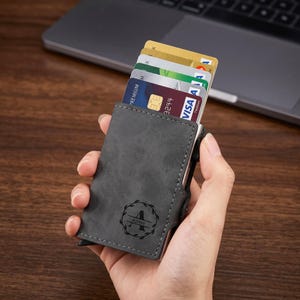 Op de afbeelding: Een grijze leren kaarthouder met een zilveren metalen frame, met daarin verschillende creditcards. De kaarthouder heeft een cirkelvormig monogramontwerp met de letter "A" en de naam "AJANI". De achtergrond omvat een laptop en een houten oppervlak.