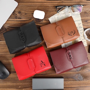 May include: Four personalized leather wallets in black, brown, red, and burgundy. Each wallet features a unique initial or monogram design. The wallets are closed with a flap and a leather strap. The wallets are on a wooden surface.