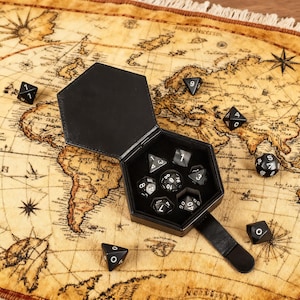 Puede incluir: Un estuche de dados hexagonal negro con una correa de cuero, abierto para revelar un conjunto de dados poliédricos negros y blancos. Varios dados están esparcidos alrededor del estuche sobre una superficie con temática de mapa. Los dados están numerados con números blancos.