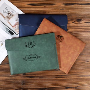 May include: Three leather document holders in navy blue, green, and tan. The green holder has the text "Chalkboard" and the initials "LR" with antlers. The tan holder has the name "William" and a mountain graphic.