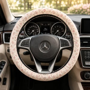 May include: A steering wheel cover featuring a light beige background with a small, repeating pink floral pattern. The cover is fitted onto a black steering wheel with a central silver Mercedes-Benz logo and integrated control buttons on the spokes.