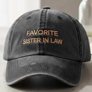 May include: A charcoal gray baseball cap with the embroidered text "FAVORITE SISTER IN LAW" in gold. The cap has a curved brim and a distressed look. The hat is sitting on a light-colored surface.
