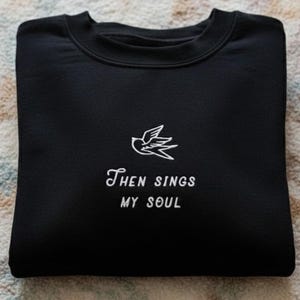 May include: A folded black sweatshirt with the embroidered phrase "Then Sings My Soul" and a bird graphic. The sweatshirt is made of a soft material and is folded neatly on a patterned surface. The design is in white.