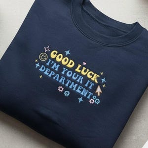 May include: Navy blue sweatshirt with the text "GOOD LUCK I'M YOUR IT DEPARTMENT" in yellow, blue, and pink. The design includes a smiley face, stars, and computer-related icons. A black coffee mug, a wooden pencil holder, and a white computer mouse are also visible.
