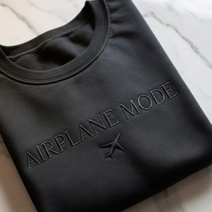 Puede incluir: Sudadera negra de cuello redondo con las palabras "AIRPLANE MODE" bordadas en el pecho, junto con un peque&ntilde;o gr&aacute;fico de avi&oacute;n. La sudadera est&aacute; doblada cuidadosamente sobre una superficie de m&aacute;rmol.