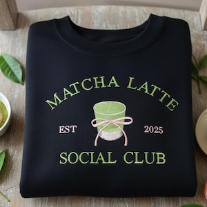 May include: Black sweatshirt with green embroidered text "Matcha Latte Social Club" and a graphic of a matcha drink. The sweatshirt is on a wooden chair, with matcha accessories and macarons.