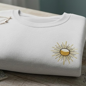 May include: A folded white sweatshirt with a crew neck and a gold sunburst design embroidered on the front. A silver necklace rests on the folded sweatshirt. The sweatshirt is on a wooden surface.