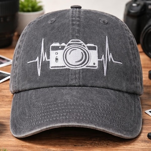 May include: A gray baseball cap featuring a white embroidered camera design with a heartbeat line. The cap has a curved brim and a classic six-panel construction. Perfect for photographers and photography enthusiasts.