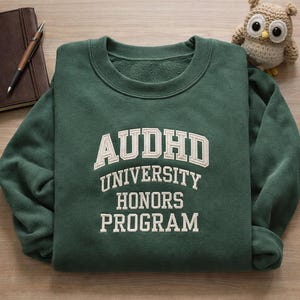 May include: A dark green sweatshirt with the text "AUDHD UNIVERSITY HONORS PROGRAM" in white. A small, crocheted owl figurine is in the upper right corner. A brown notebook and pen are in the upper left corner.