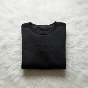 Puede incluir: Sudadera negra de cuello redondo con las palabras "AIRPLANE MODE" y un peque&ntilde;o gr&aacute;fico de avi&oacute;n bordado en la parte delantera. La sudadera est&aacute; doblada cuidadosamente sobre una superficie blanca y esponjosa.