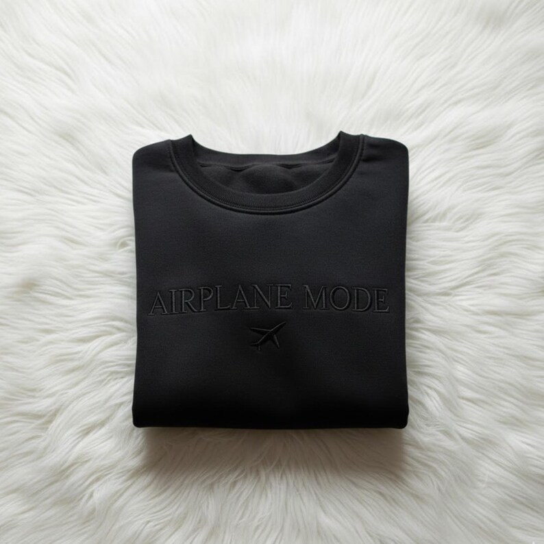 Puede incluir: Sudadera negra de cuello redondo con las palabras "AIRPLANE MODE" y un peque&ntilde;o gr&aacute;fico de avi&oacute;n bordado en la parte delantera. La sudadera est&aacute; doblada cuidadosamente sobre una superficie blanca y esponjosa.
