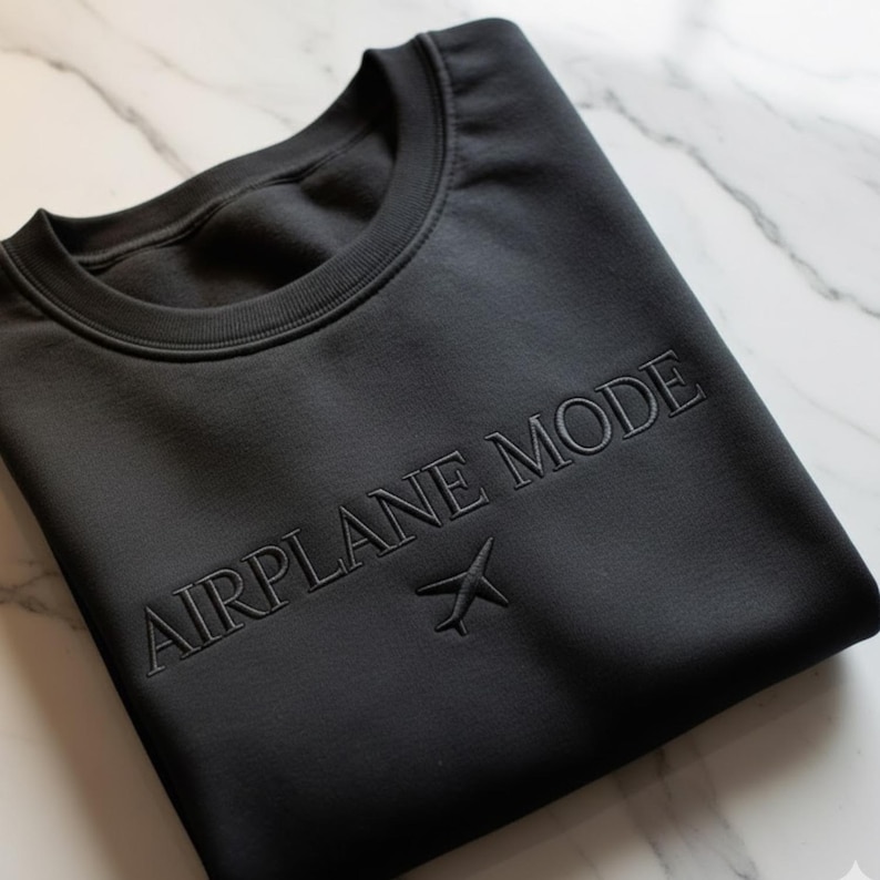 Puede incluir: Sudadera negra de cuello redondo con las palabras "AIRPLANE MODE" bordadas en el pecho, junto con un peque&ntilde;o gr&aacute;fico de avi&oacute;n. La sudadera est&aacute; doblada cuidadosamente sobre una superficie de m&aacute;rmol.