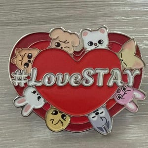Puede incluir: Pin de esmalte en forma de corazón rojo con el texto "#LoveSTAY" en blanco. El pin presenta personajes de animales de dibujos animados alrededor del corazón. El pin tiene un borde plateado y está sobre un fondo gris claro.