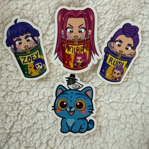 May include: Four stickers on a white, textured surface. Three stickers depict anime-style characters in ramen cups, with names like "Zoey" and "Rumi." A blue cat sticker with large yellow eyes and a hat is also present.