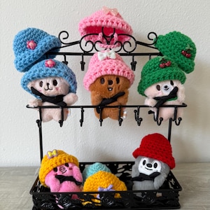 May include: A black metal display stand holds several plush bear figures, each wearing a different colored crocheted hat. The hats are in shades of blue, pink, green, yellow, and red. The bears have black mustaches and bows.