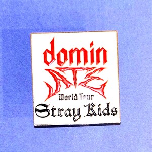 May include: A square pin with a white background and a red and black design. The pin features the words "domin" and "ATEZ" in a stylized font, with "World Tour Stray Kids" below.