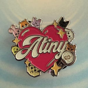 Op de afbeelding: Decoratieve pin met een roze hart met het woord "Atiny" in witte letters. Het hart is omgeven door cartoon dierenfiguren, sterren en een microfoon. De pin heeft een zilveren rand.