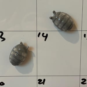 May include: Two gray ceramic pill bug figurines. Each bug has a segmented body and small antennae. The figurines are on a white surface with black numbers, likely a calendar or whiteboard.