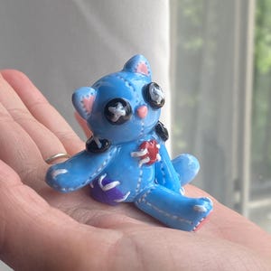 Op de afbeelding: Een kleine, lichtblauwe teddybeerfiguur met zwarte knoopogen en een rood hart. De beer heeft gestikte details en paarse accenten. Het wordt in de hand van een persoon gehouden.