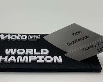 以下が含まれることがあります： 黒とシルバーのMoto GPワールドチャンピオンのトロフィー。ベースは黒で、「Moto GP WORLD CHAMPION」と書かれた白い文字があります。シルバーのプレートには「Fabio Quartararo Yamaha #20 2021」と刻まれています。