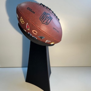 May include: A brown leather football with the NFL logo and various team logos on it, displayed on a black stand.