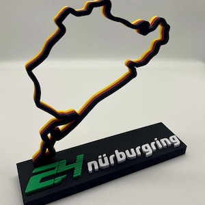Puede incluir: Un contorno en capas de una pista de carreras en negro, amarillo y rojo, montado sobre una base negra. La base muestra la palabra "nürburgring" en blanco y un logotipo verde. El contorno de la pista es una representación estilizada.