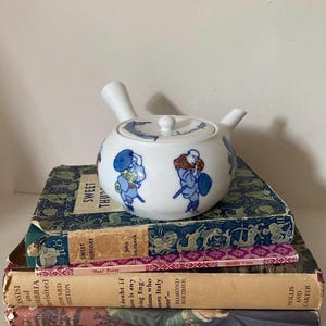 May include: A white ceramic teapot featuring blue and white illustrations of Japanese figures. The teapot is placed on a stack of vintage books with patterned covers. The teapot has a handle and a spout.