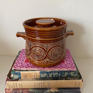 May include: A brown ceramic casserole dish with a lid and handles, featuring a decorative swirl pattern. The dish is stacked on top of a pile of vintage books with colorful covers. The casserole dish is a kitchenware item.