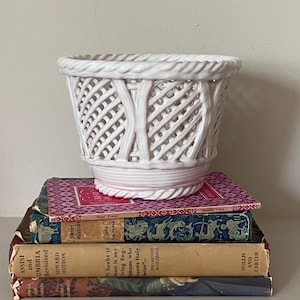 May include: A white ceramic planter with a woven basket design, placed on a stack of vintage books. The planter features a braided rim and a lattice pattern. The books have colourful covers and titles.