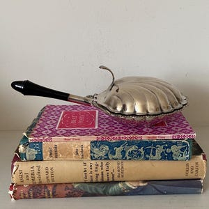 May include: A silver clam shell-shaped serving dish with a black handle sits on a stack of vintage books. The books have colourful covers and titles including "Beat Poets" and "Sweet Thursday". The dish is a decorative item.