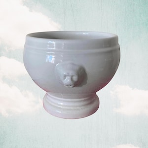 May include: White ceramic soup tureen with a lion head design on the side. The tureen has a round body and a pedestal base.