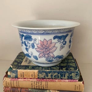 May include: A white ceramic bowl featuring blue floral and leaf patterns and a pink lotus flower design. The bowl rests on a stack of vintage books with gold lettering and decorative covers. The bowl is approximately 15 cm in diameter.