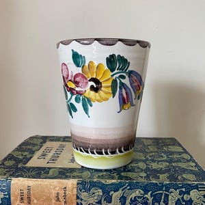 May include: A white ceramic mug with a floral design. The flowers are painted in shades of yellow, pink, blue, and green. The mug has a brown rim and a brown band around the bottom.