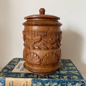May include: A wooden jar with a lid, intricately carved with floral and bird designs. The jar is brown and has a smooth, polished finish.