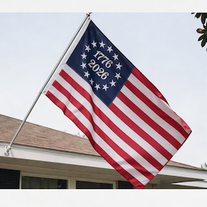 May include: A flag with red and white stripes and a blue canton with white stars and the numbers "1776" and "2026". The flag is attached to a silver pole and is displayed outside a building.