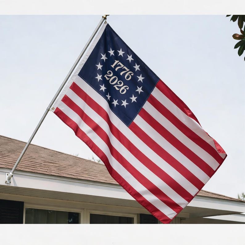 May include: A flag with red and white stripes and a blue canton with white stars and the numbers "1776" and "2026". The flag is attached to a silver pole and is displayed outside a building.
