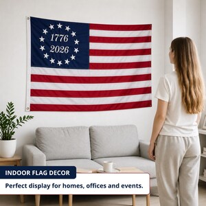 May include: An American flag with red and white stripes and a blue section with white stars and the numbers 1776 and 2026. The flag is displayed on a white wall in a living room.