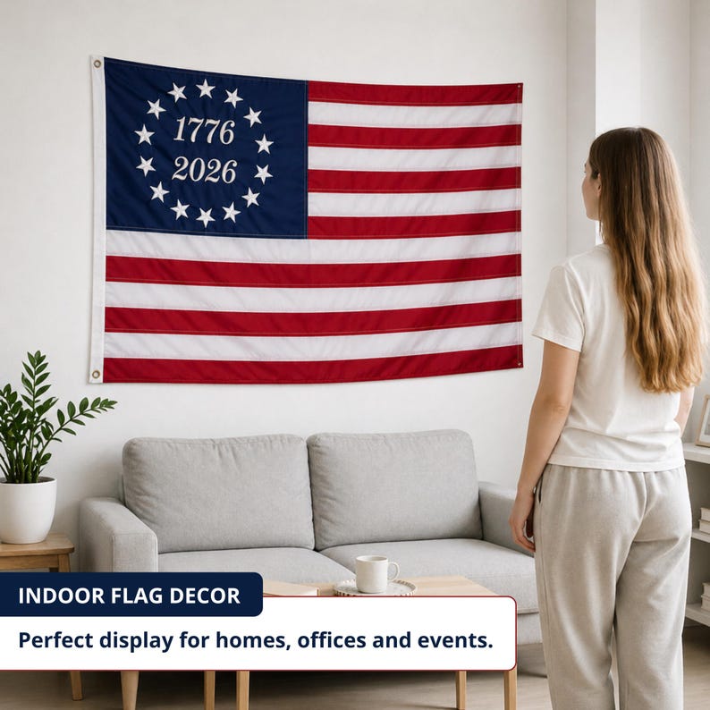 May include: An American flag with red and white stripes and a blue section with white stars and the numbers 1776 and 2026. The flag is displayed on a white wall in a living room.