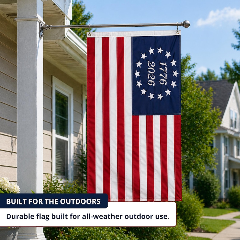 May include: A vertical flag with red and white stripes and a blue canton featuring 13 white stars in a circle, with the numbers "1776" and "2026" inside. The flag is displayed outdoors, attached to a silver pole.