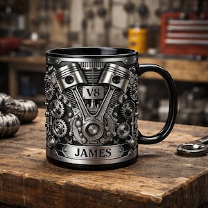 Könnte beinhalten: Schwarze Kaffeetasse mit detailliertem silbernen Motordesign, einschließlich Kolben, Zahnrädern und dem Text "V8". Die Tasse hat einen schwarzen Griff und der Name "JAMES" ist auf der Vorderseite zu sehen. Auf einer Holzoberfläche.