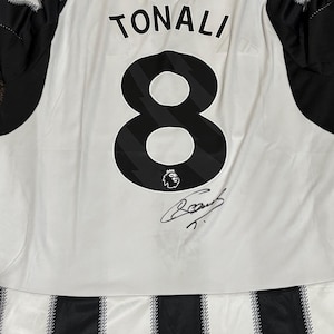 May include: White and black soccer jersey with the name "TONALI" and the number "8" printed on the back. The jersey has a signature and the words "HOWAY THE LADS" printed on the top.