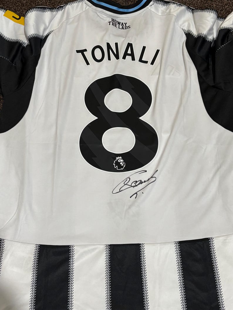 May include: White and black soccer jersey with the name "TONALI" and the number "8" printed on the back. The jersey has a signature and the words "HOWAY THE LADS" printed on the top.