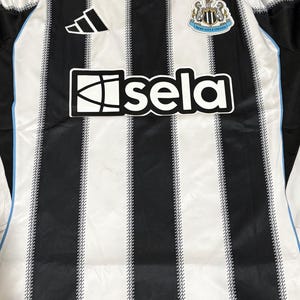 May include: A black and white striped soccer jersey with the word "isela" across the front. The jersey has a black collar and the Adidas logo on the upper left. The Newcastle United crest is on the upper right.