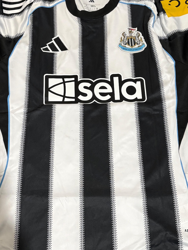 May include: A black and white striped soccer jersey with the word "isela" across the front. The jersey has a black collar and the Adidas logo on the upper left. The Newcastle United crest is on the upper right.