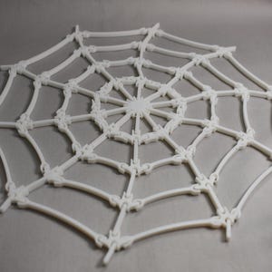 May include: A white, three-dimensional spider web, constructed from interconnected, curved lines. The web's design features a central hub with radiating spokes and concentric circles, creating a detailed and symmetrical pattern. The web is set against a neutral background.